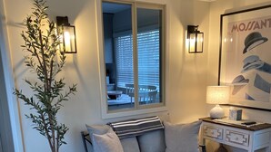Ett modernt vardagsrum med en soffa, en pall och stolar, en krukväxt, ett fönster med persienner och en vägglampa.