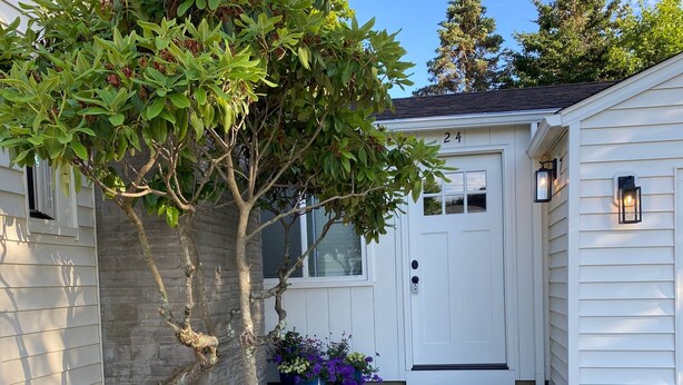 Ett vitt hus med en ytterdörr, ett träd och en blomsterrabatt med rosa blommor.