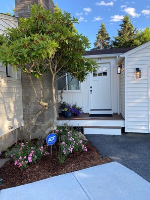 Une maison blanche avec une porte d’entrée, un arbre et un parterre de fleurs roses.