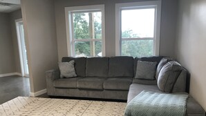 Un salon avec un canapé d’angle gris, un tapis à motifs et une grande fenêtre donnant sur des arbres.