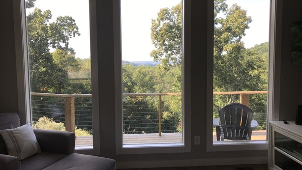 Une pièce avec un canapé gris, un tapis à motifs et une vue sur un balcon agrémenté d’une chaise et d’une balustrade en bois.