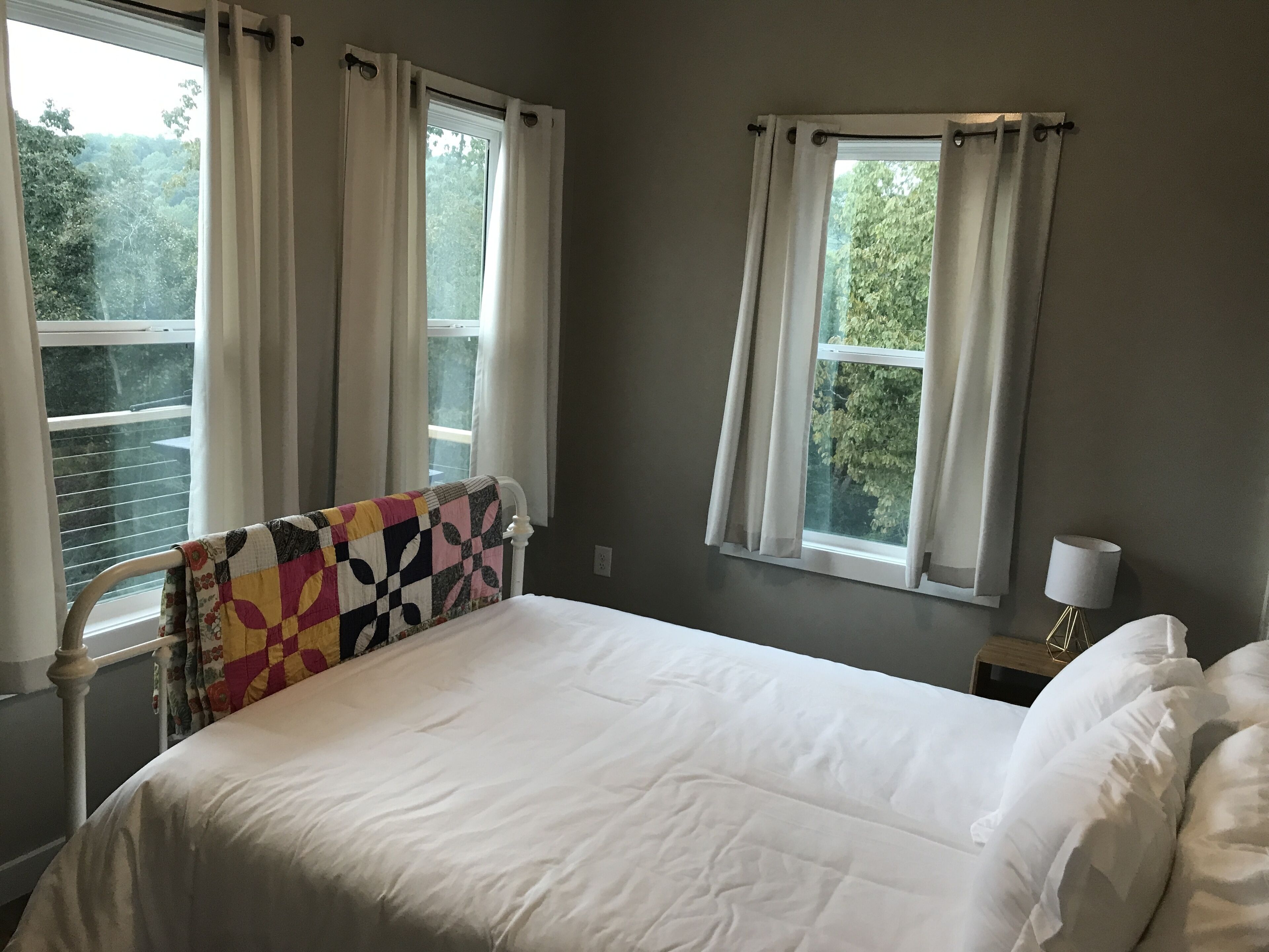 A bedroom with a bed, two windows with curtains, a bedside table with a lamp, and a view of trees outside.