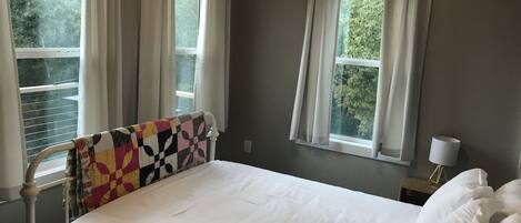 A bedroom with a bed, two windows with curtains, a bedside table with a lamp, and a view of trees outside.