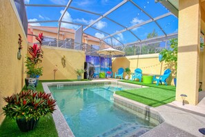 Un área de piscina con techo de vidrio, césped artificial y sillas azules.