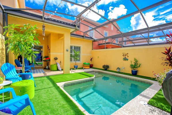 Una piscina con techo de vidrio, rodeada de paredes amarillo brillante, sillas azules y césped artificial verde.