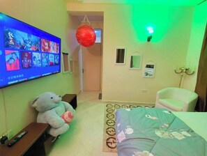 A hotel room with a TV displaying a program, a teddy bear, a bed with a floral bedspread, a white armchair, and a red spherical decoration.
