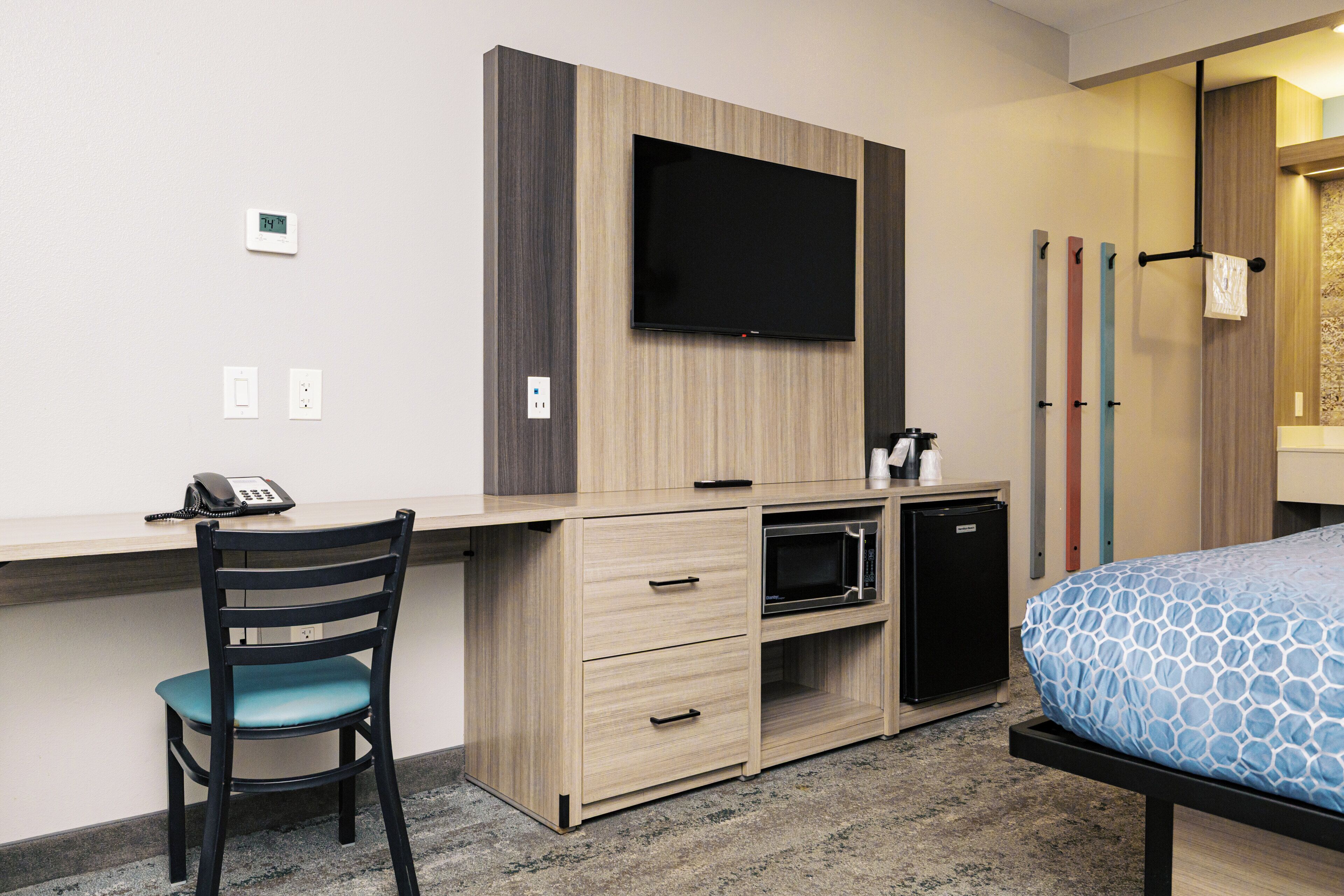 A hotel room with a desk, chair, bed, television, and a small kitchenette.