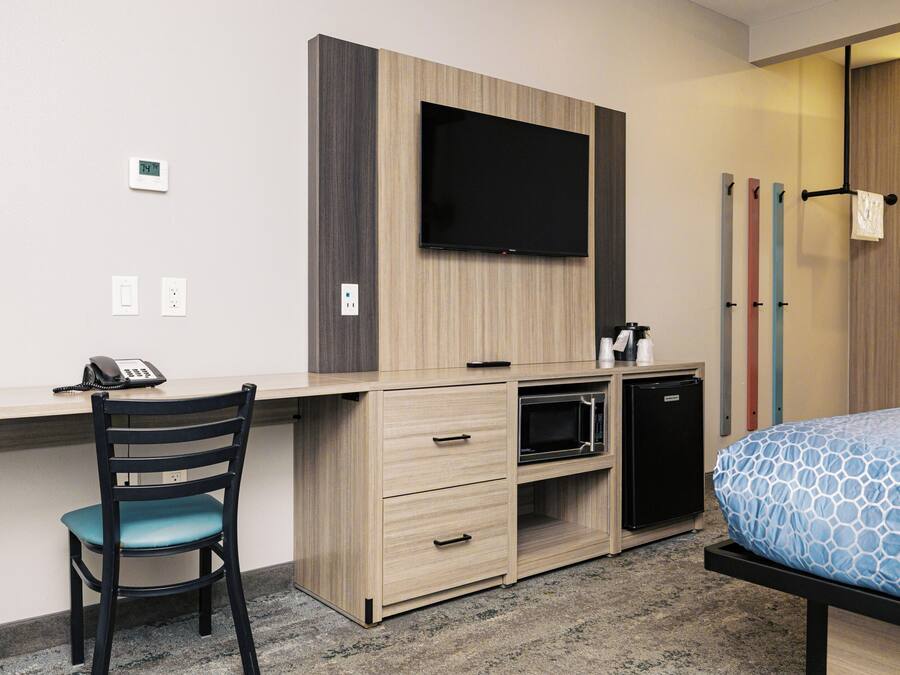A hotel room with a desk, chair, bed, television, and a small kitchenette.
