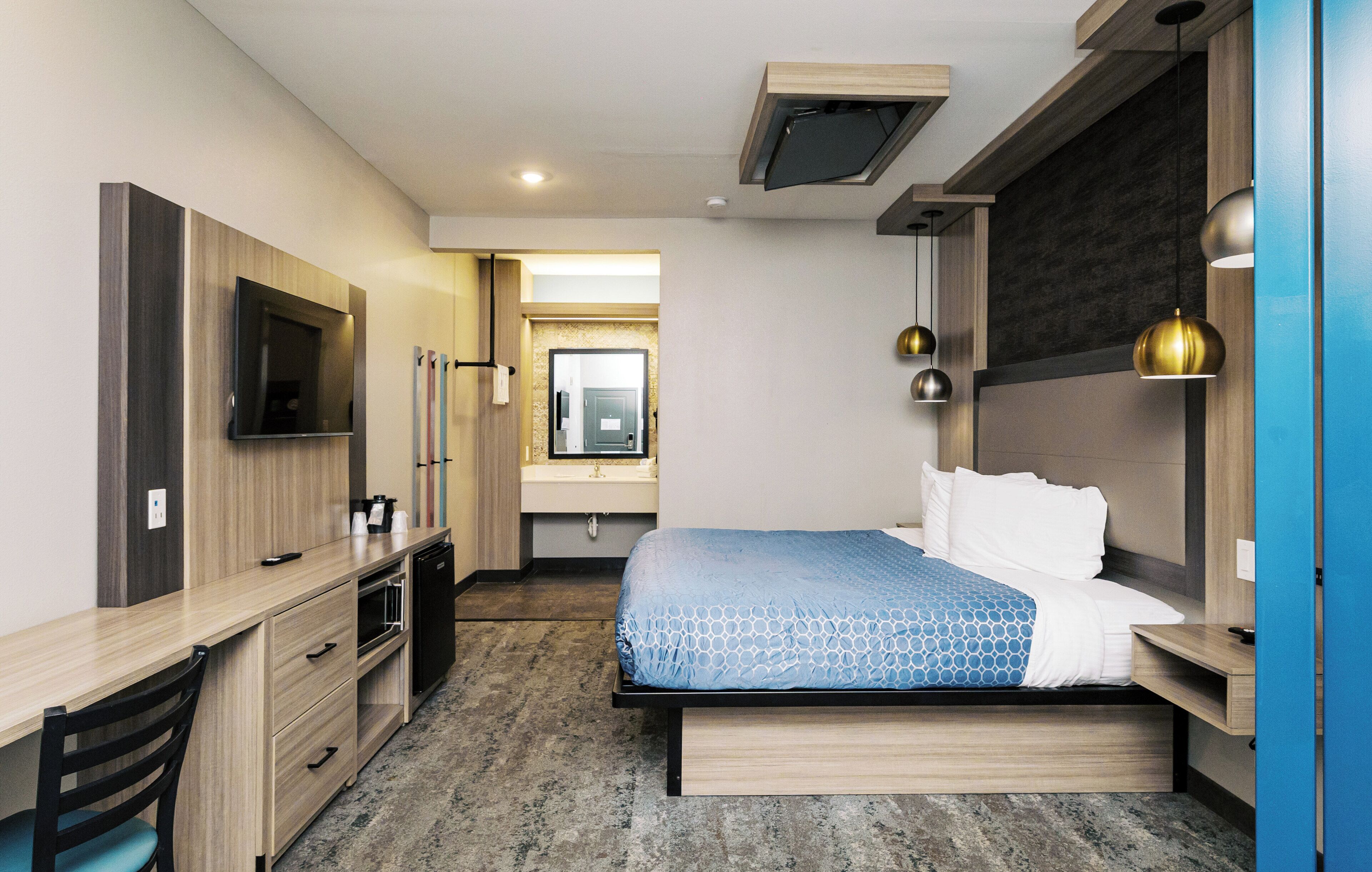 Une chambre d’hôtel moderne équipée d’un lit, d’un bureau, d’une télévision et d’une salle de bain visible à travers une porte ouverte.