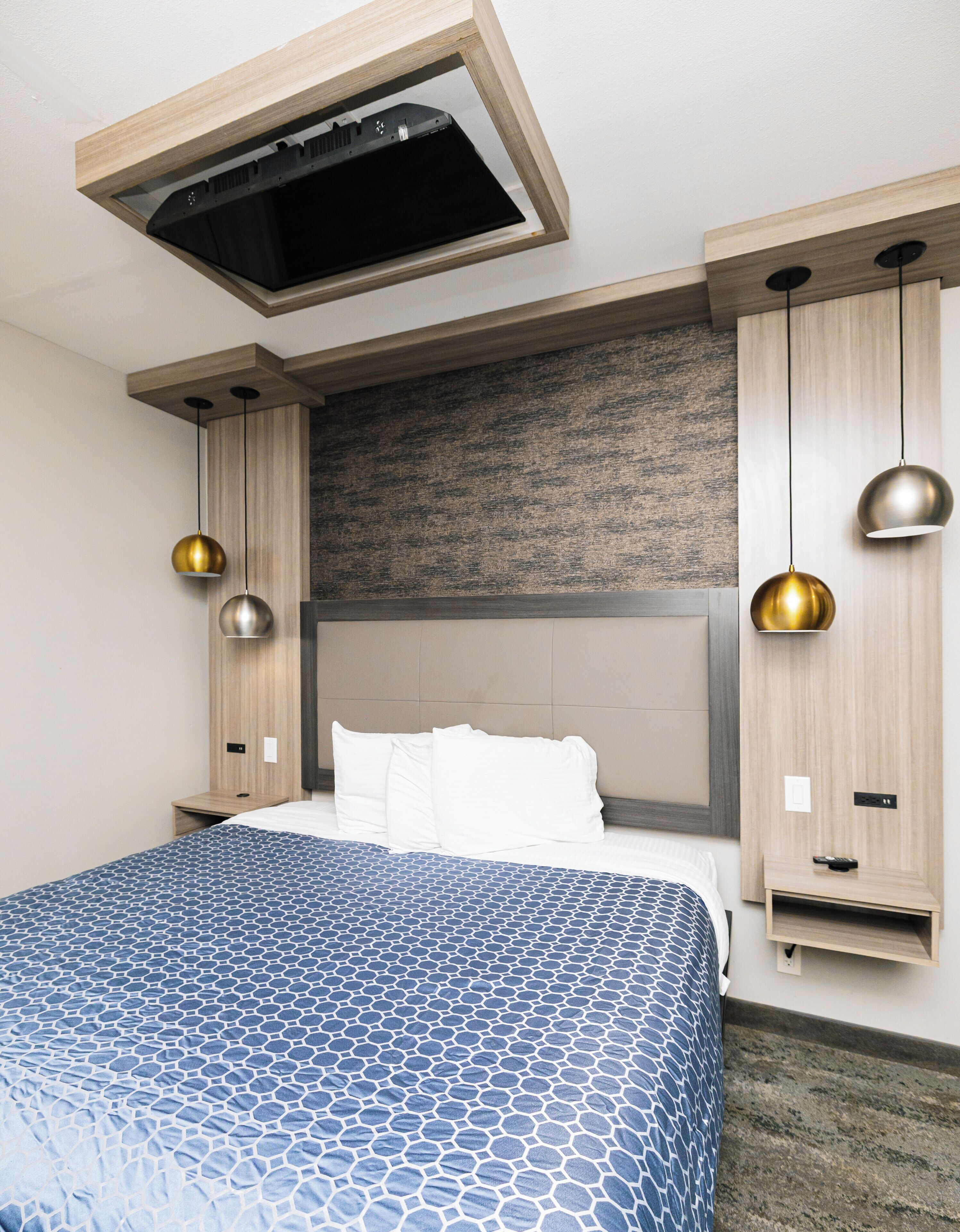 A hotel room with a large bed, a built-in headboard, pendant lighting, and a wall-mounted control panel.