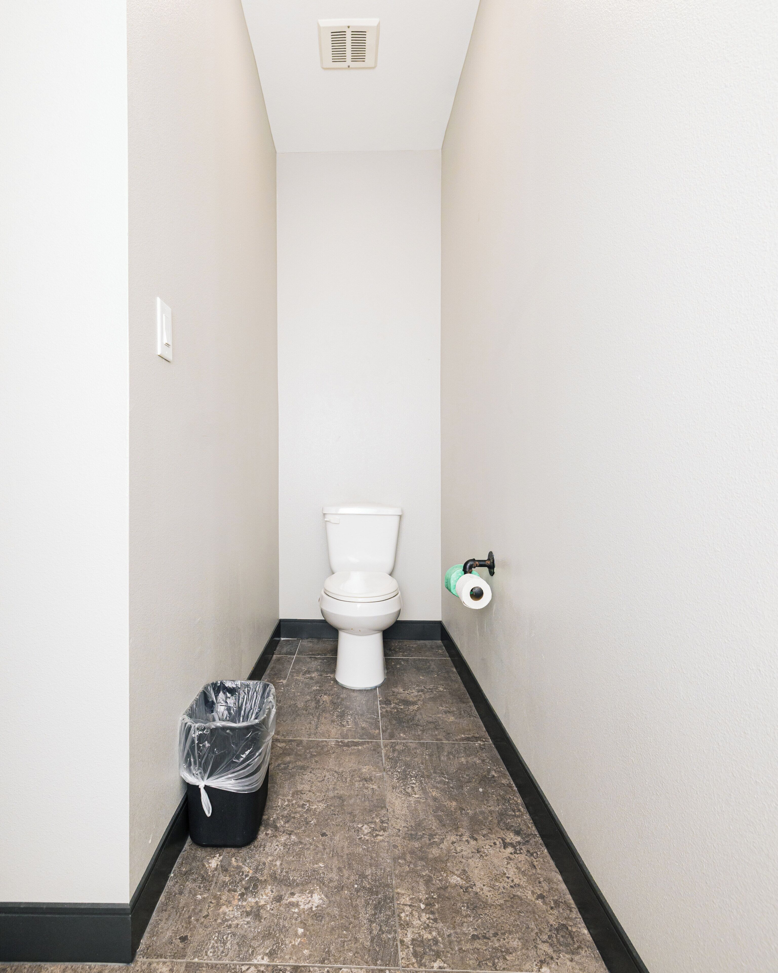A narrow hallway with a white toilet, a trash bin, and a toilet paper holder.