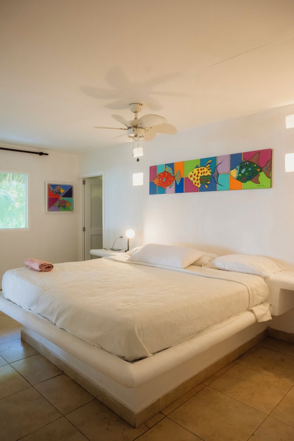 A bedroom with a bed, a ceiling fan, a nightstand with a lamp, and a colorful fish painting on the wall.