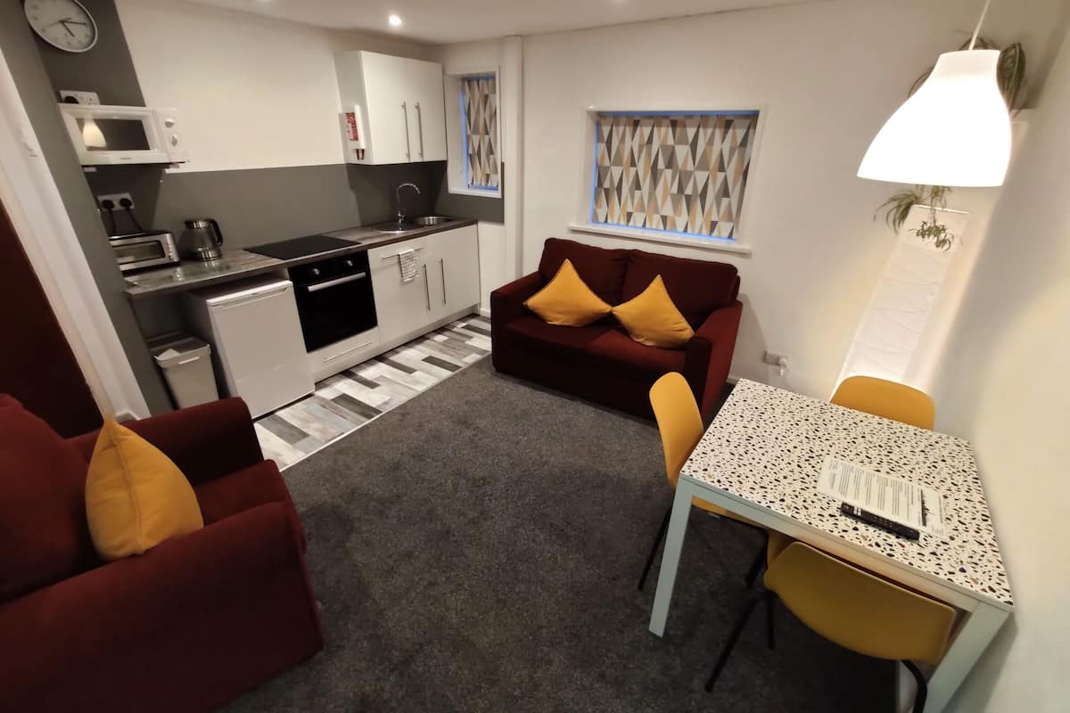 A compact living space with a kitchenette, a sofa, a dining table, and a small window with geometric curtains.