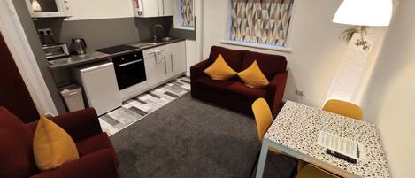 A compact living space with a kitchenette, a sofa, a dining table, and a small window with geometric curtains.