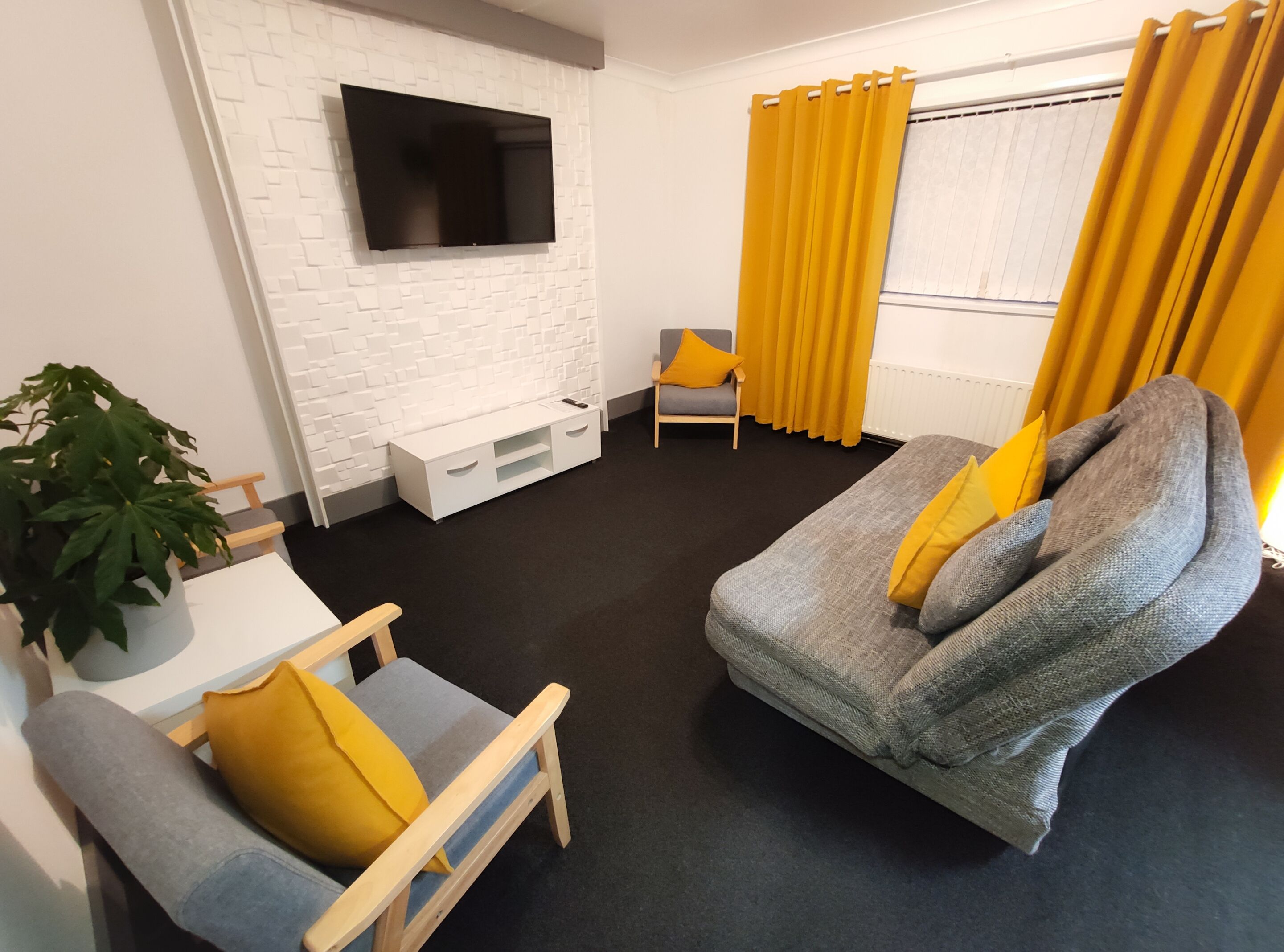 A modern living room with a flat-screen TV mounted on a white brick wall, a gray sofa with yellow cushions, a wooden chair, a small white table, and a potted plant.