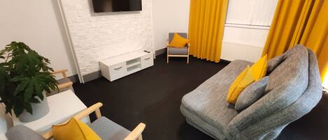 A modern living room with a flat-screen TV mounted on a white brick wall, a gray sofa with yellow cushions, a wooden chair, a small white table, and a potted plant.