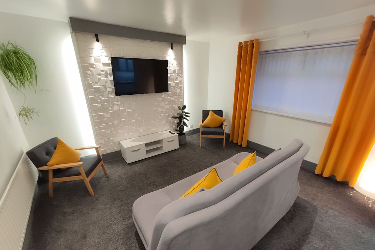 A modern living room with a grey sofa, two armchairs, a white entertainment unit, and a flat-screen TV mounted on a textured wall.