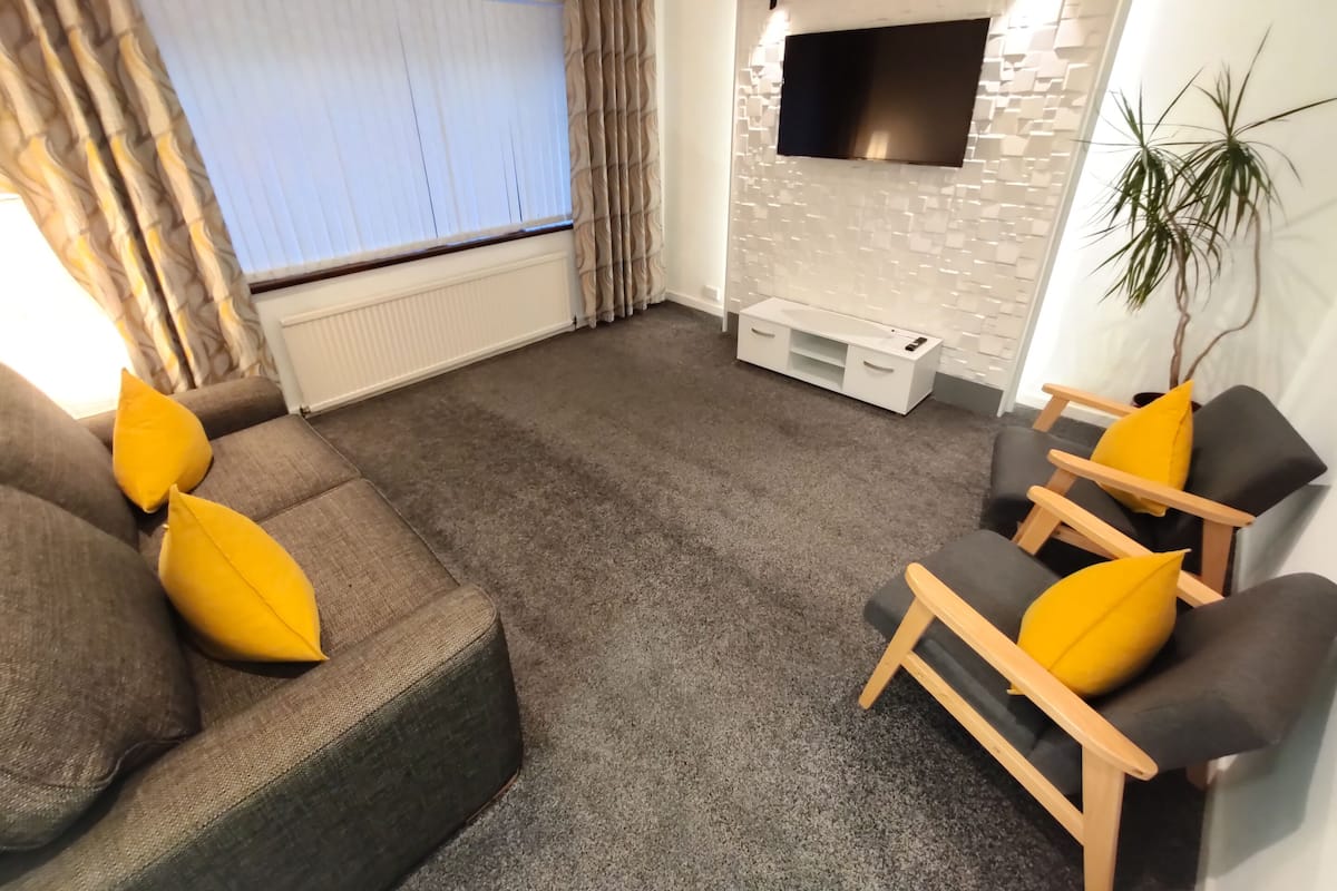 A modern living room with a grey carpet, a grey sofa with yellow cushions, a wooden chair with a yellow cushion, a white entertainment unit, and a flat-screen TV mounted on a white brick wall.