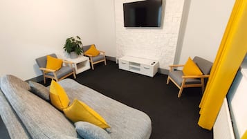 Una sala de estar moderna con un sofá gris, dos sillones, un mueble blanco para equipos electrónicos y un televisor de pantalla plana montado sobre una pared de ladrillo blanco.