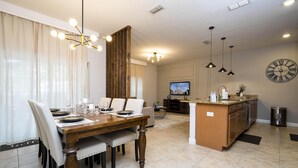 A modern dining area with a wooden table and chairs, a kitchen with a built-in island, and a large wall clock. - 4873- Great Family Home near Disney, private pool, Storey Lake (Pet Friendly) (Kissimmee)