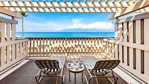 A balcony with two chairs and a table, overlooking a beach with clear blue water and a palm tree. - Double Lanai, Double Ocean Views, all comfort (Lahaina)