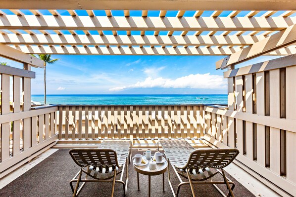 A balcony with two chairs and a table, overlooking a beach with clear blue water and a palm tree.