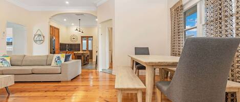 A spacious living room with a wooden floor, a grey sofa, a dining table with chairs, and a kitchen in the background.