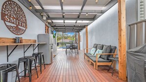 Een overdekt buitengebied met een houten terras, een zithoek met een bank en stoelen, een bar met krukken, en een groot decoratief bord aan de muur.