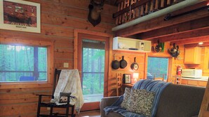 A cozy cabin interior with a wooden floor, a sofa, a rocking chair, a dining table, and a kitchen area with hanging pots. - Beyersfort next to Cygnet Lake - Sleeps 5, Private Dock w/ Canoe, No Wi-Fi (Condon)
