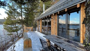 A wooden cabin with large windows overlooking a lake, a wooden deck, and a bench.