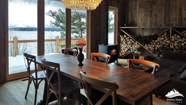 A wooden interior with a large dining table, chairs, a chandelier, and a view of a lake through large windows.