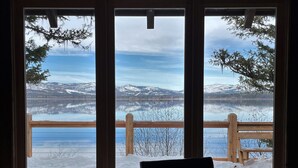 A room with a view of a snowy landscape through large windows, a wooden deck outside, and a black chair inside.