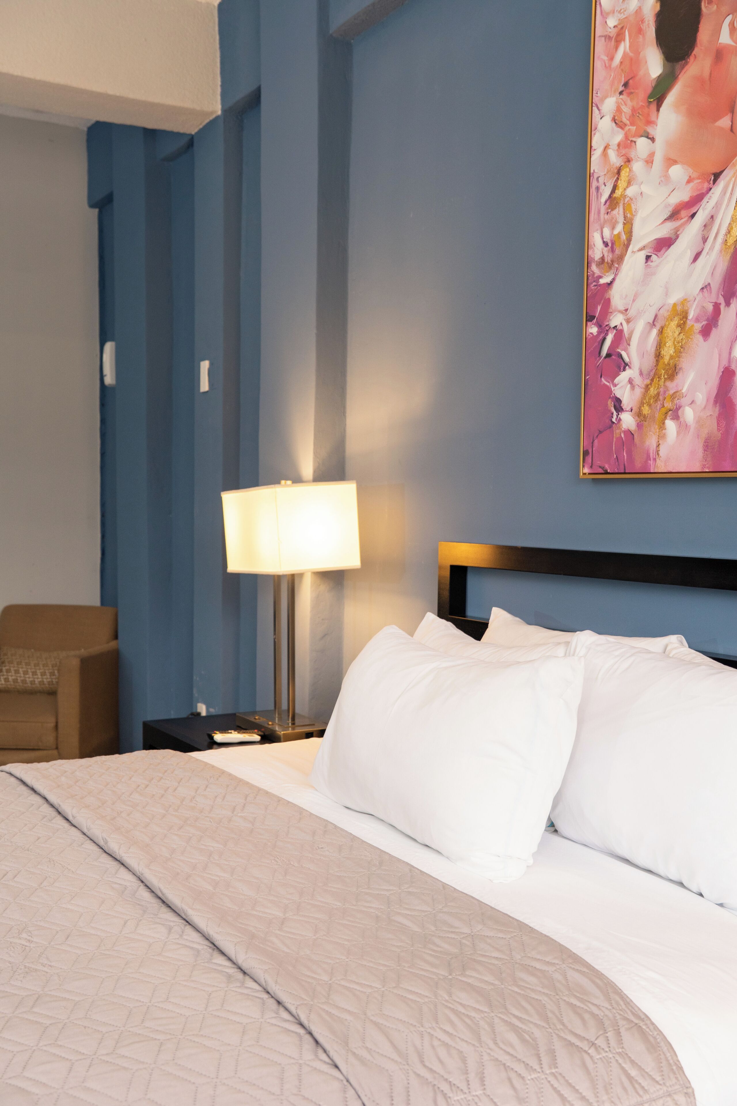 Habitación de hotel con una cama, una mesita de noche con lámpara, una silla y un cuadro abstracto grande en la pared.