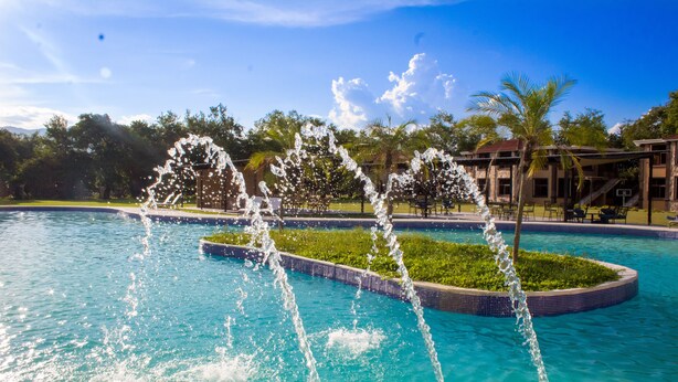 A swimming pool with water jets, surrounded by palm trees and a building with a basketball court. - Vista Terra (Chiquimula)