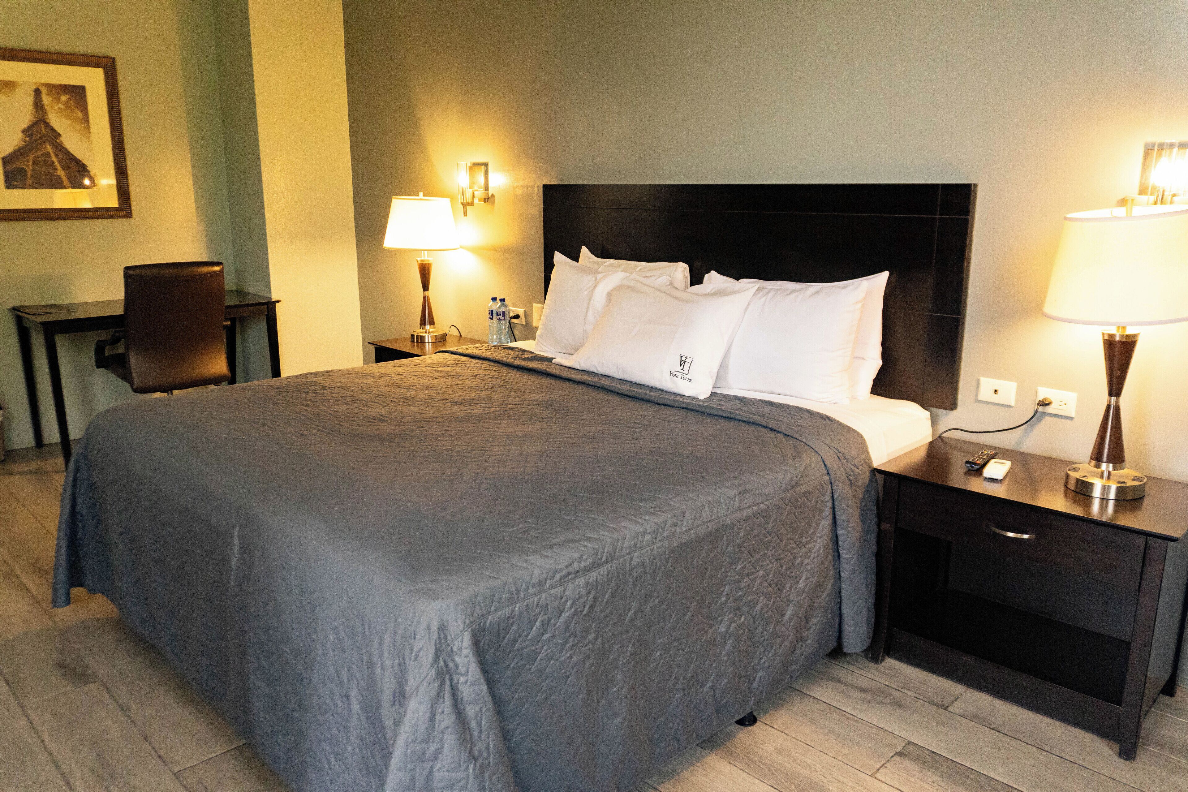 A neatly made bed with a grey bedspread, two bedside tables with lamps, a chair, and a framed picture on the wall.
