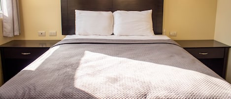 Una cama bien tendida con un cabecero oscuro, dos mesitas de noche con lámparas y una ventana con cortinas.