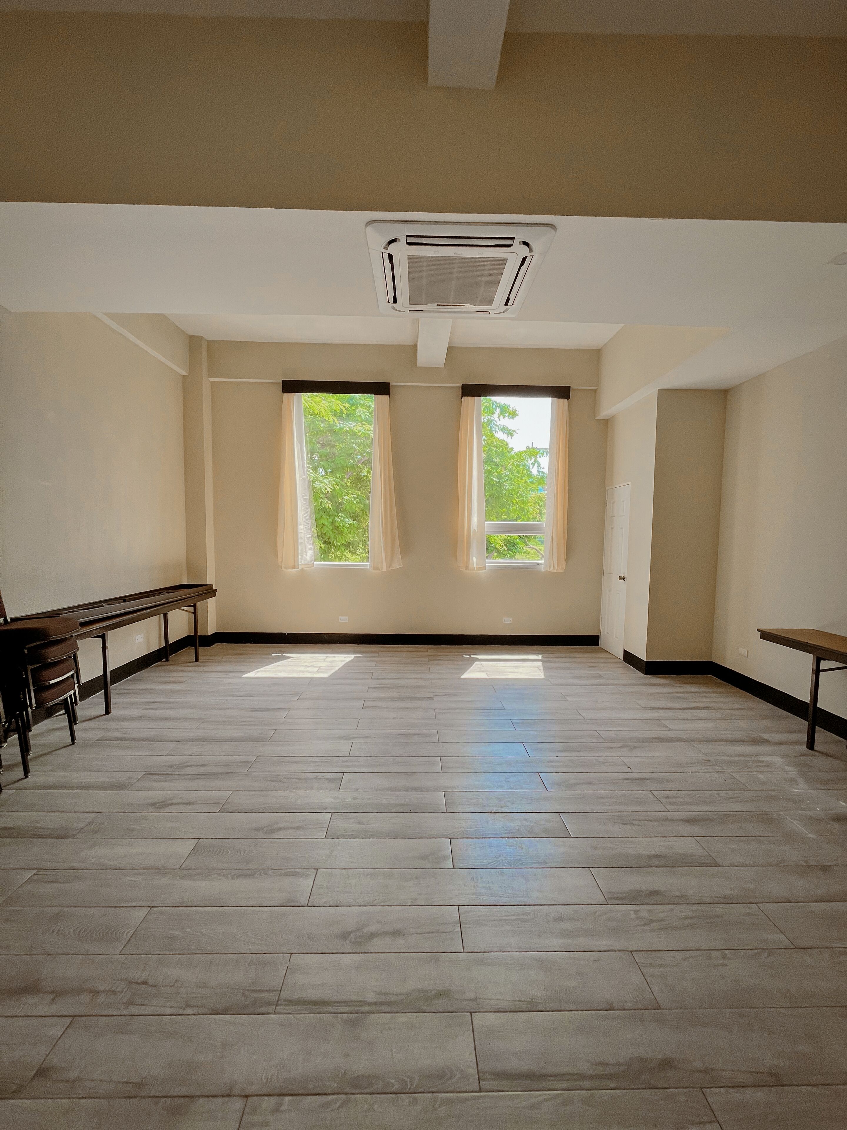 A spacious room with a herringbone floor pattern, two windows with curtains, a ceiling-mounted air conditioning unit, and a bench along one wall.