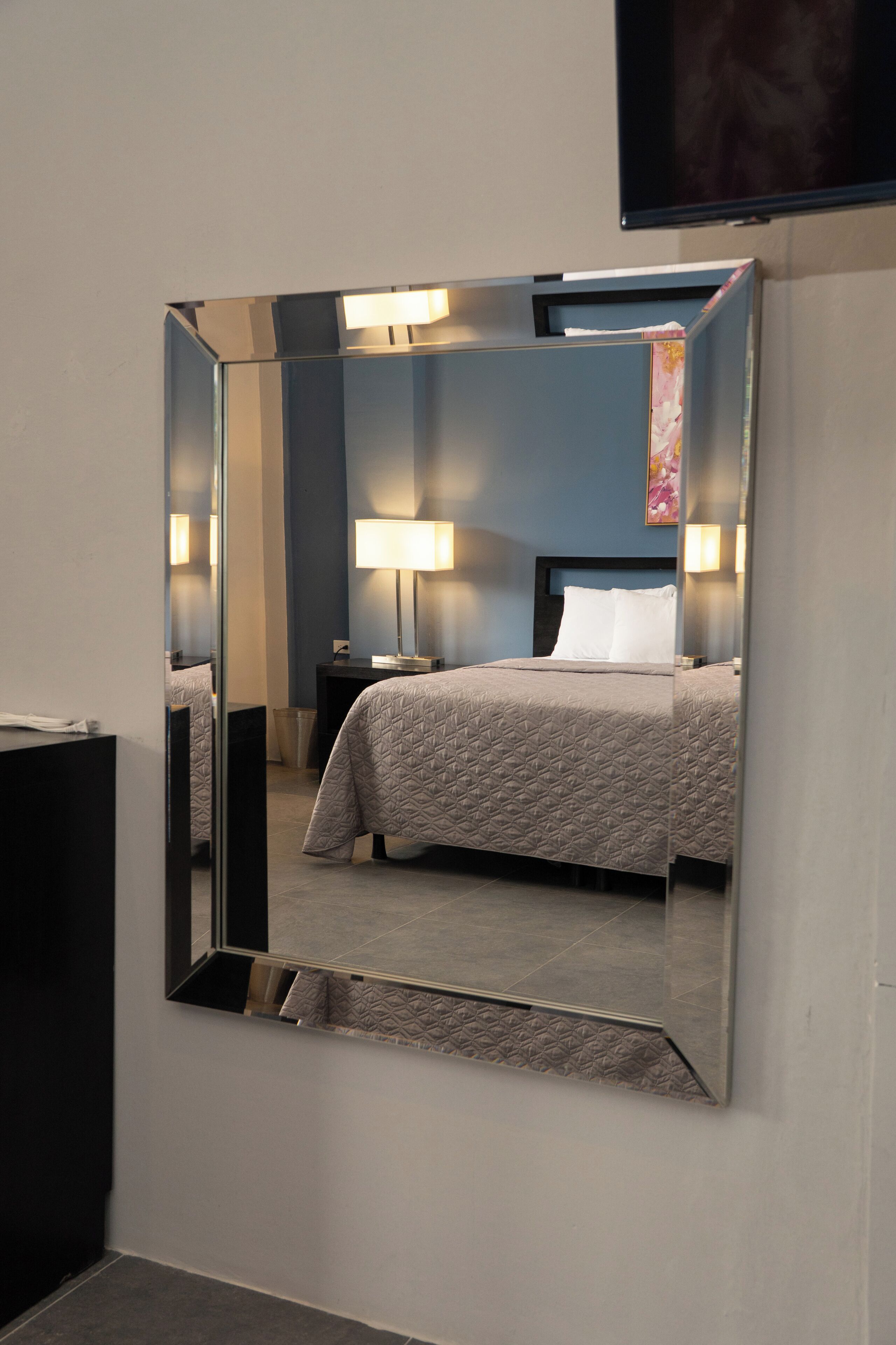 A hotel room with a large mirror, a bed with a patterned bedspread, bedside lamps, and a flat-screen TV mounted on the wall.