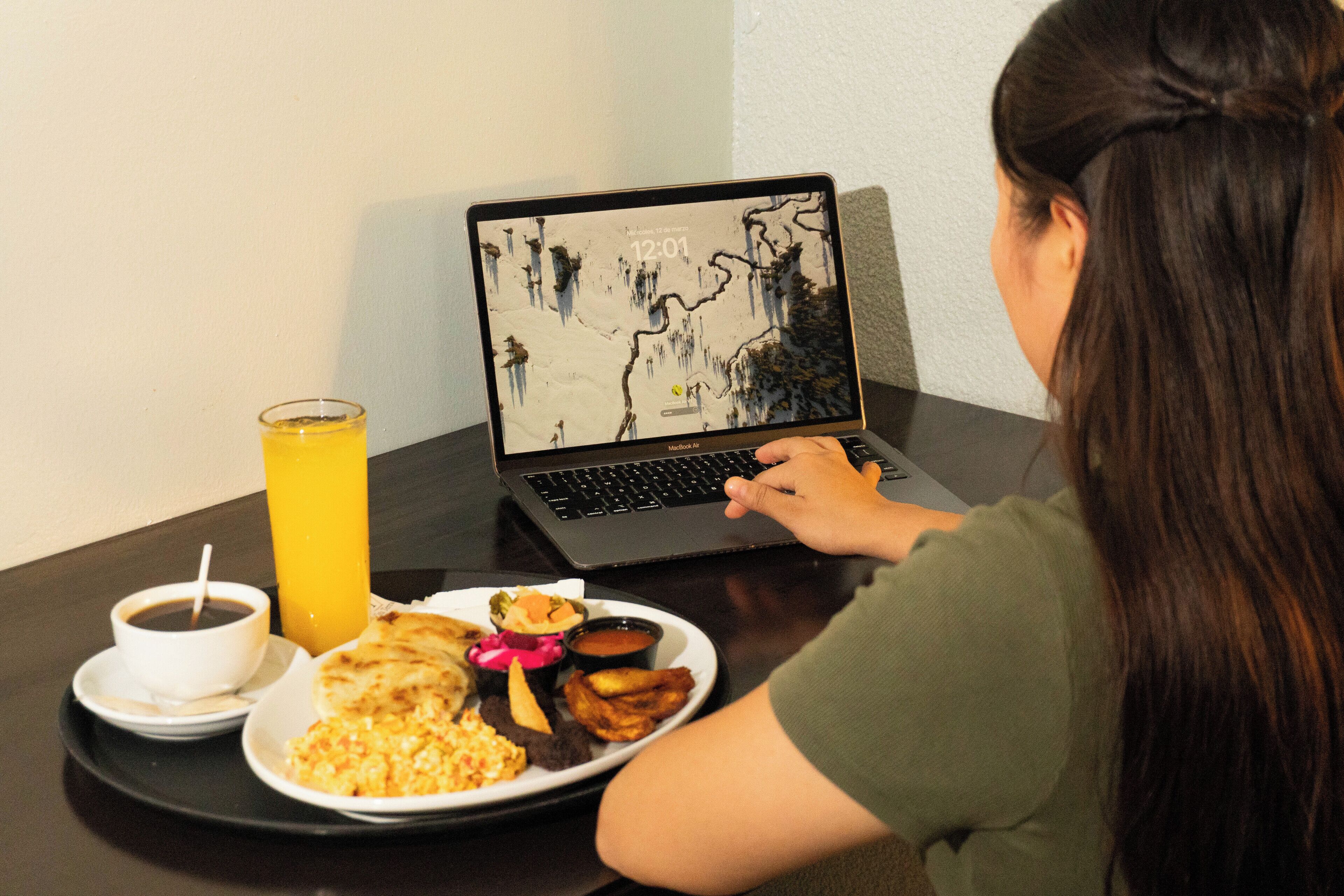 A person using a laptop at a table with a meal consisting of a plate with various dishes, a glass of orange juice, and a cup of coffee.