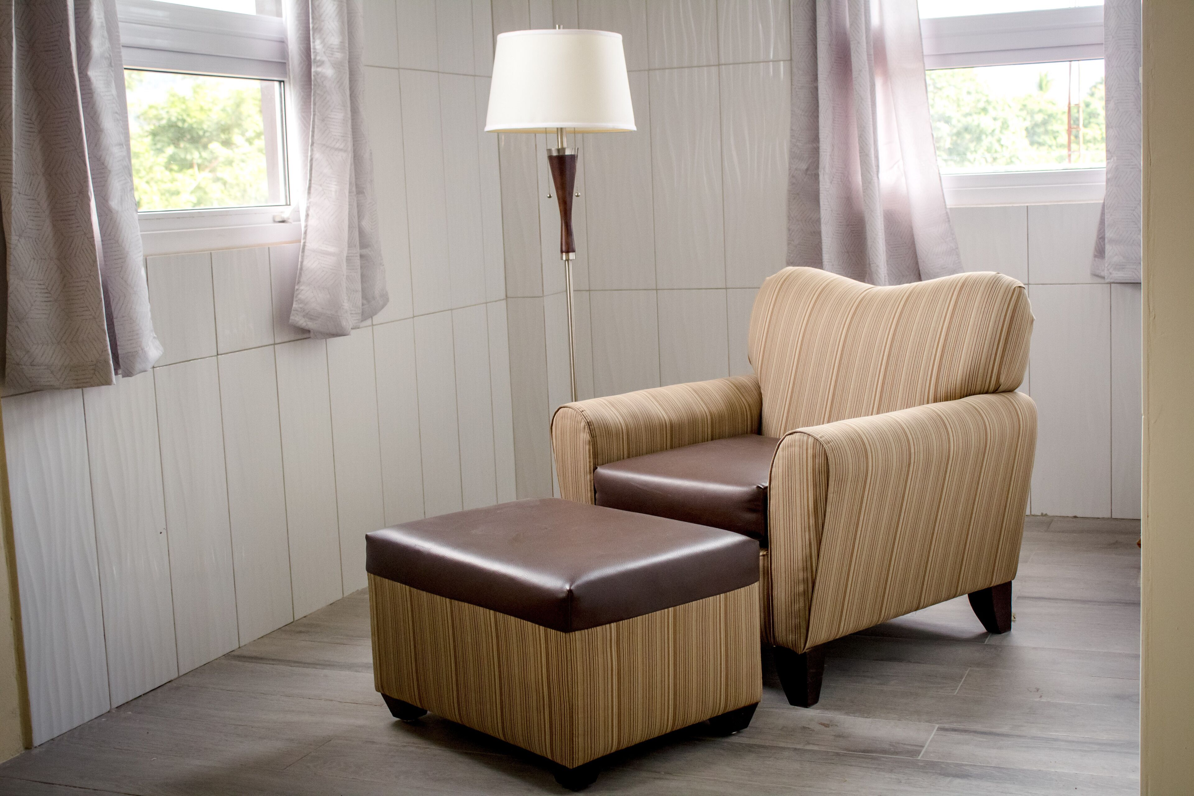 Ein Zimmer mit einem beigen Sessel, dazu passendem Fußschemel, einer Stehlampe und einem Fenster mit Vorhängen.