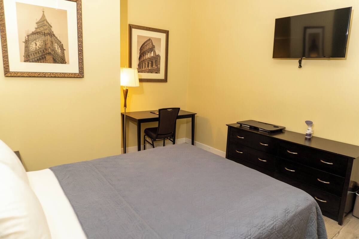 A hotel room with a bed, a desk, a chair, a dresser, and a television mounted on the wall.