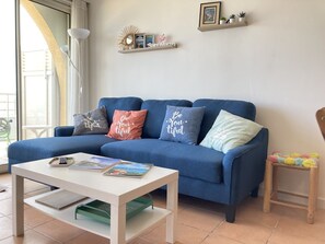 En stue med en blå sofa, et hvidt sofabord og en lille træstol. Der er dekorative puder på sofaen og en reol med billeder på væggen.