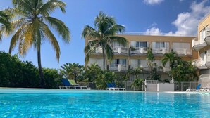 A clear blue pool with lounge chairs and palm trees, surrounded by a multi-story building with balconies.