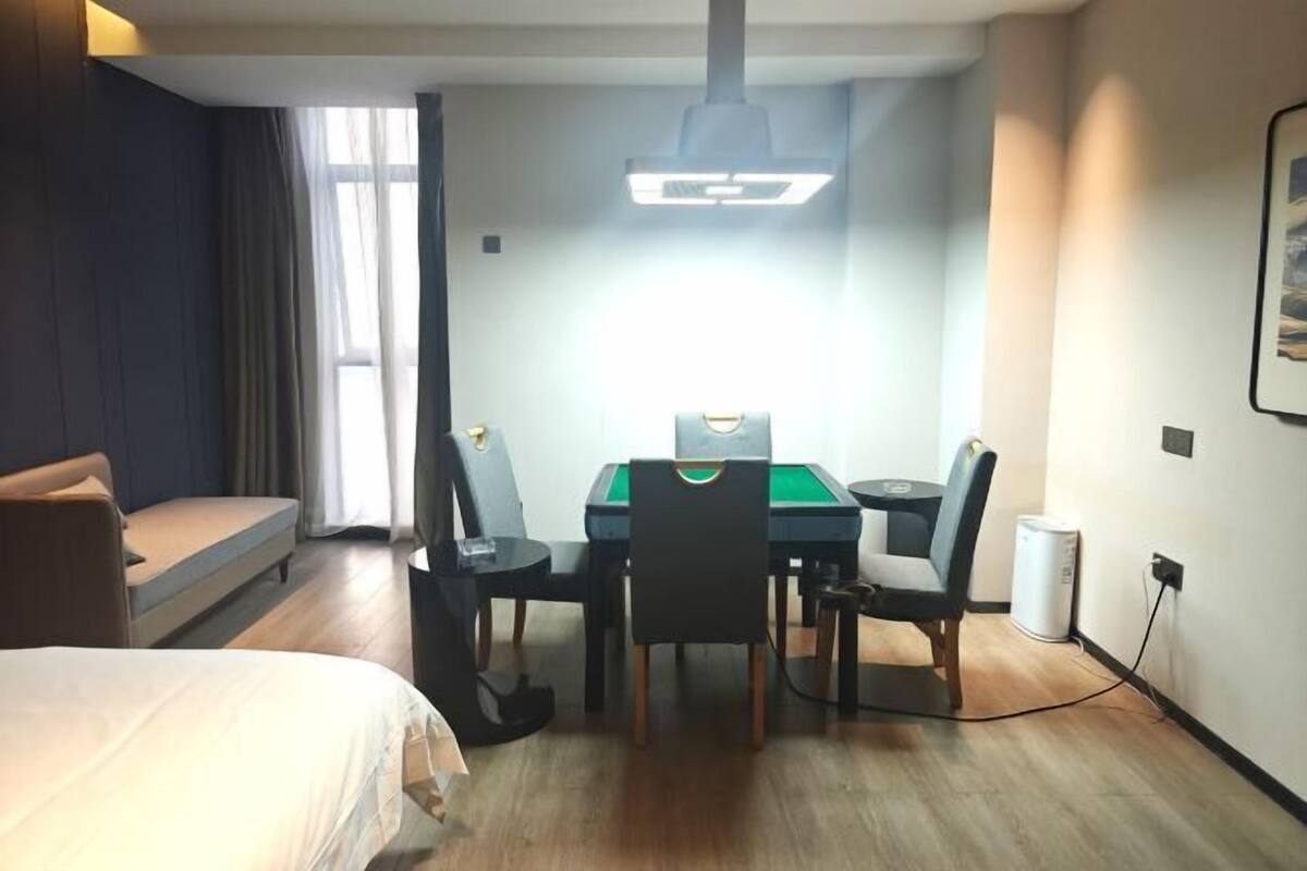 Una habitación de hotel con una cama, un escritorio con mantel verde, dos sillas, una lámpara y una ventana con cortinas.