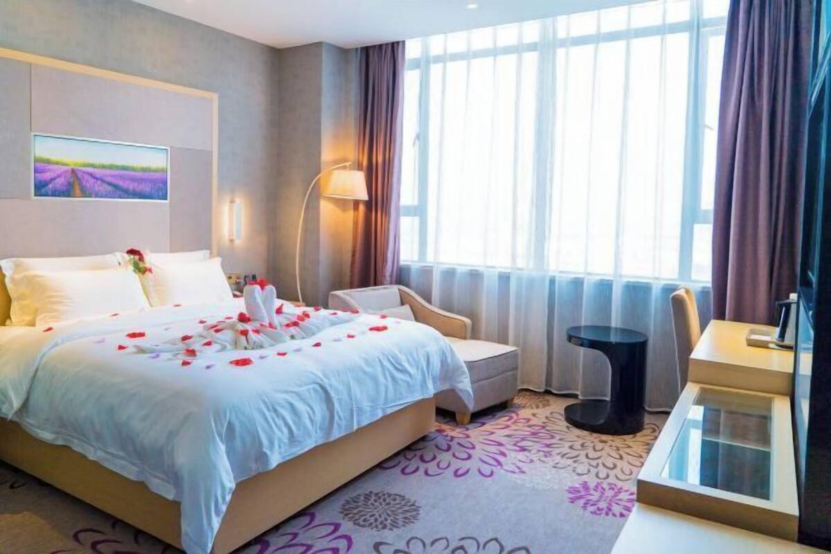 Una habitación de hotel con cama, sillón, mesita de noche, lámpara y ventana con cortinas.