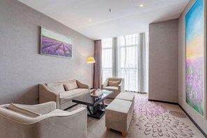 A modern hotel room with a sofa, armchair, ottoman, and a glass coffee table. There is a large window with curtains, a painting on the wall, and a patterned rug on the floor. - Lavande Hotel (Huizhou Yuanzhou) (Boluo)