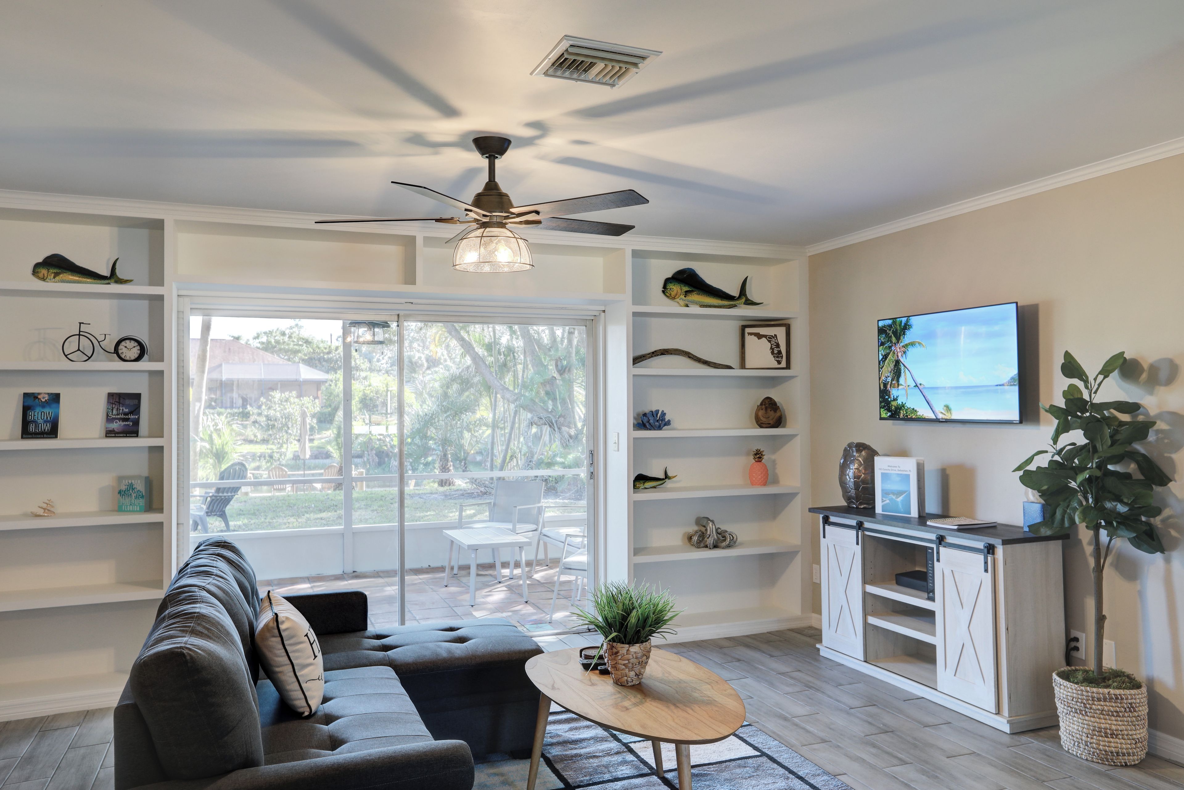A modern living room with a sofa, a coffee table, and a TV on a wall. There are shelves with decorative items and a ceiling fan.