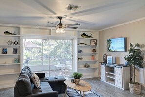 A modern living room with a sofa, a coffee table, and a TV on a wall. There are shelves with decorative items and a ceiling fan.