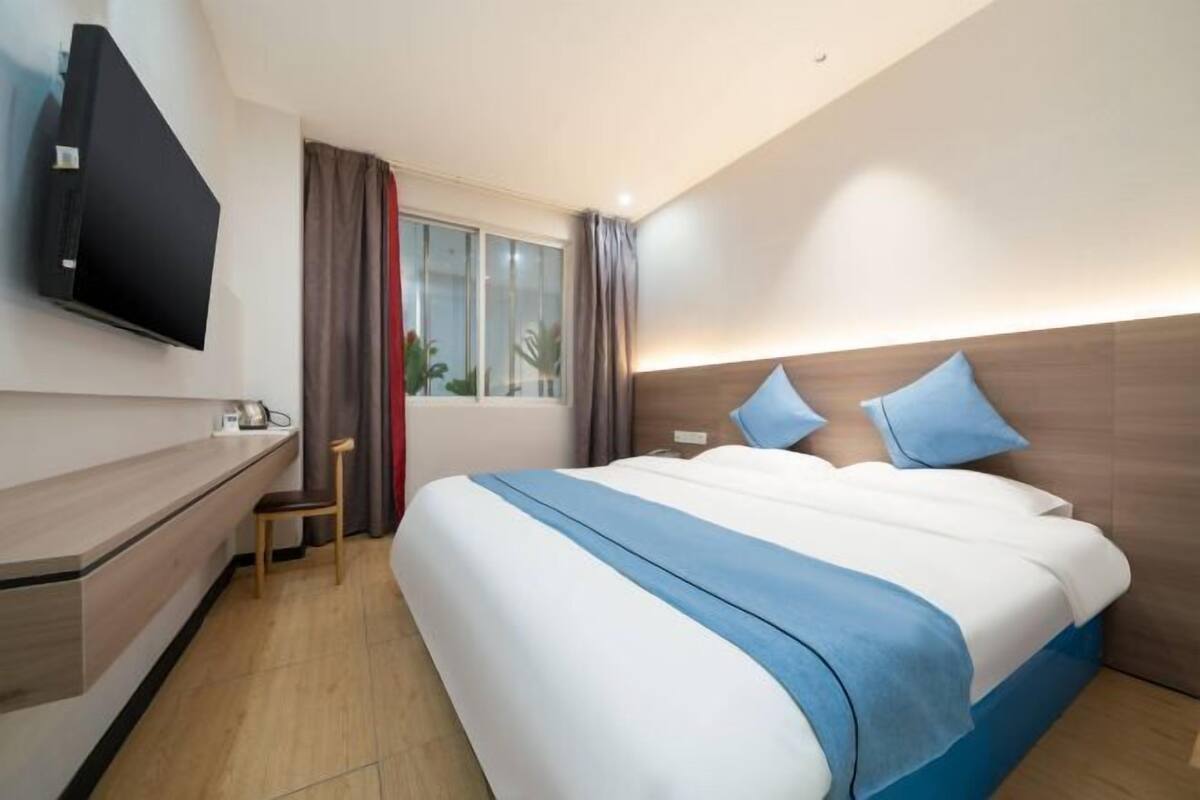 A hotel room with a bed, a TV mounted on the wall, a desk, and a window with curtains.