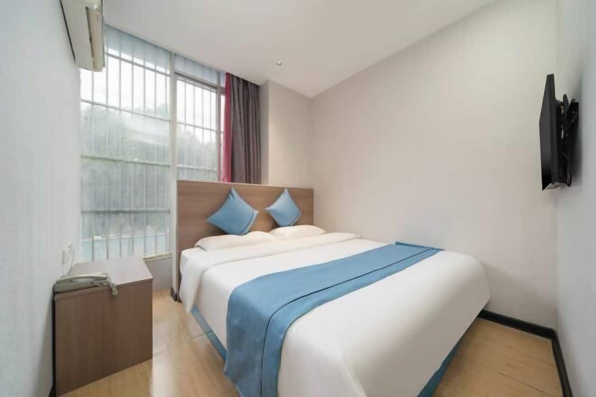 A hotel room with a bed, a desk, a chair, a window with curtains, and a wall-mounted TV.